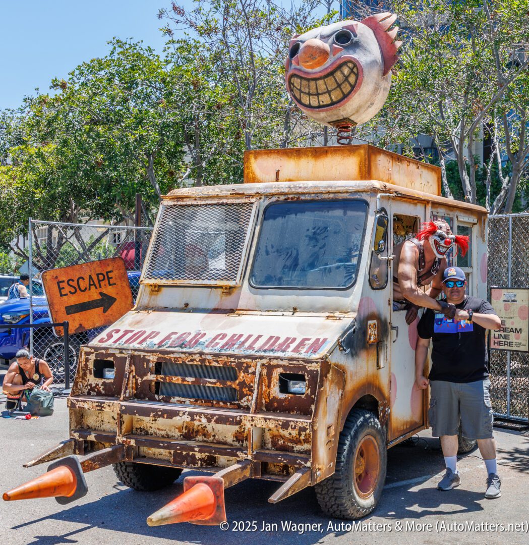 A rusty, modified truck with clown decorations and spikes is parked outdoors; a costumed person poses with another person in front of an "ESCAPE" sign.