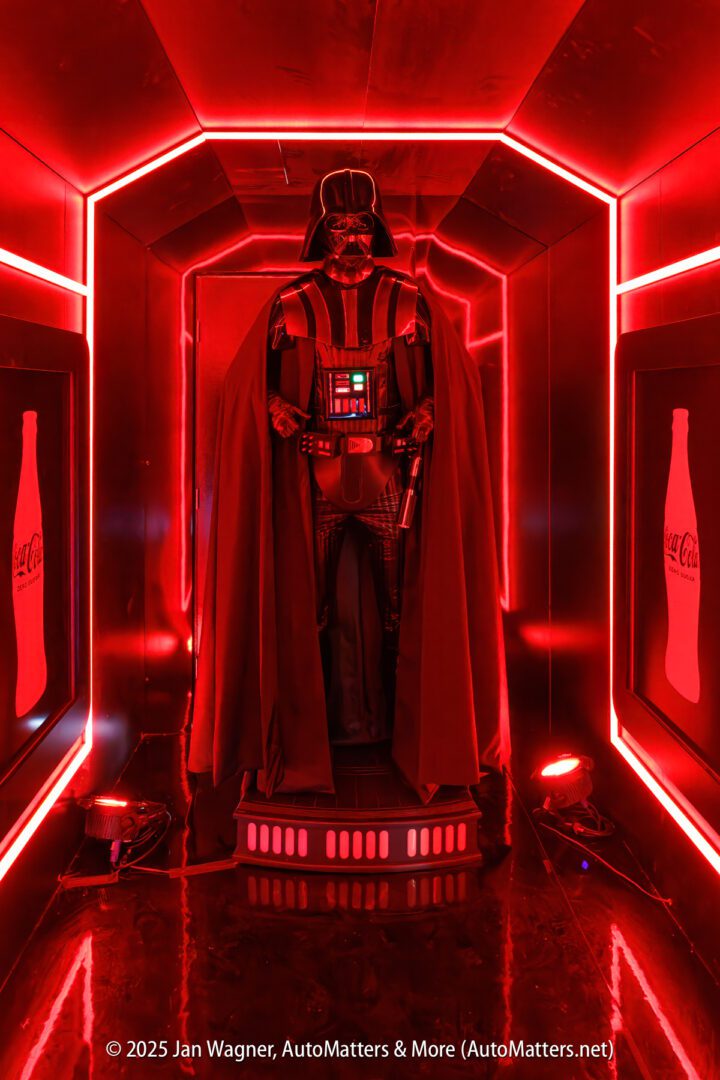 A person dressed as Darth Vader stands in a red-lit corridor with Coca-Cola logos on the walls.