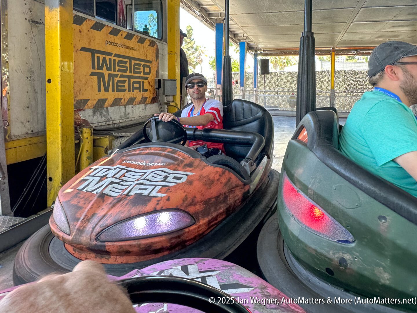 Two men are riding bumper cars themed with "Twisted Metal" branding at an amusement park.