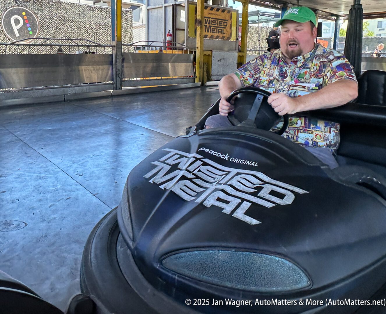 A man in a colorful shirt and green cap drives a black bumper car labeled "Twisted Metal" inside an amusement park ride area.