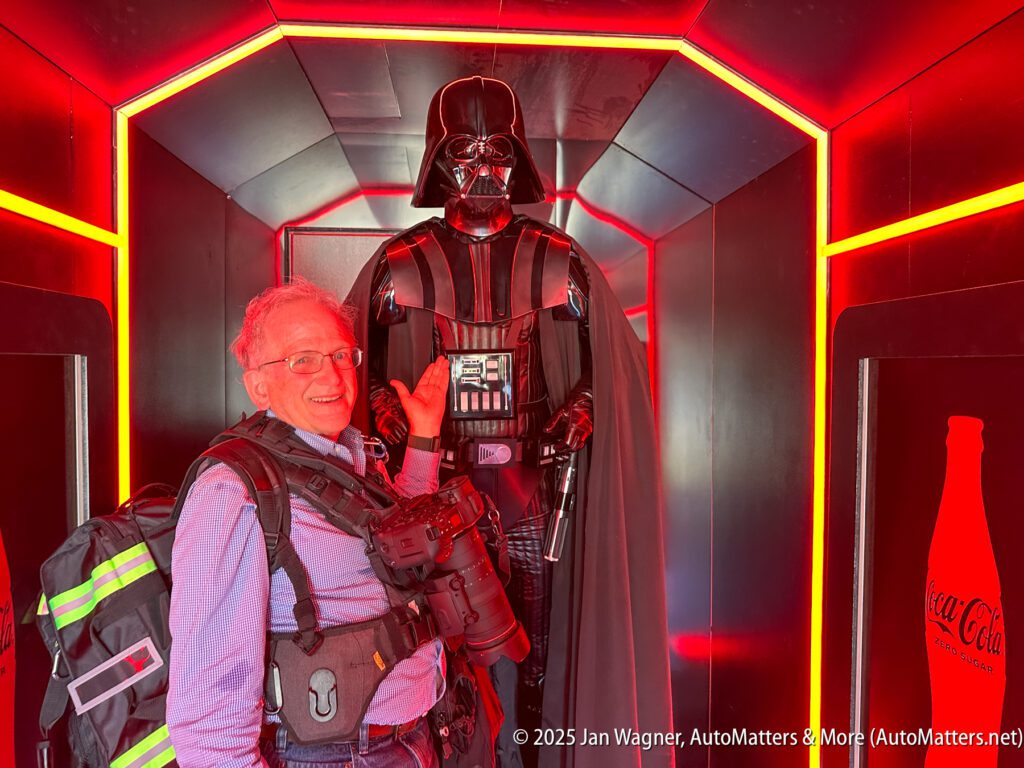 A man with camera gear stands smiling beside a Darth Vader statue in a red-lit display area.