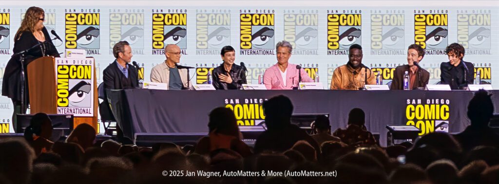 A panel of seven people sits at a long table on stage at San Diego Comic-Con International, with a moderator standing at a podium. Comic-Con banners are displayed behind them.