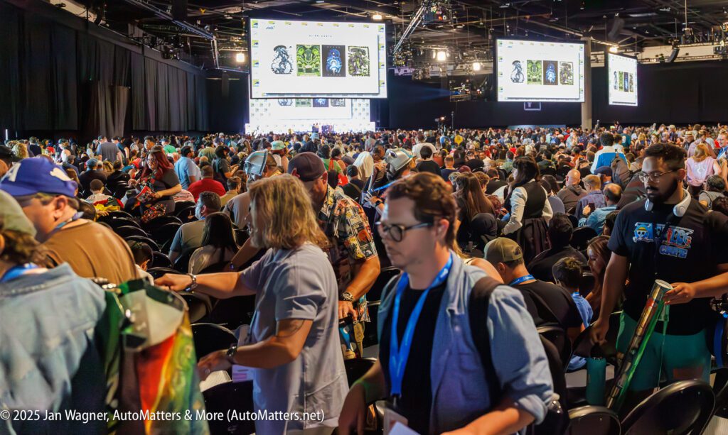 A large crowd of people stands and walks among rows of seats at a convention hall with large screens displaying images at the front.