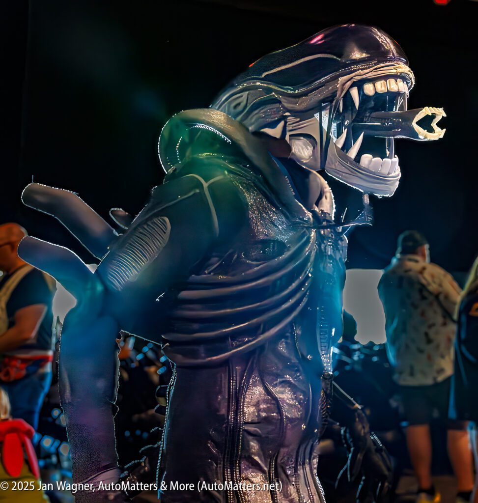 A person wears a detailed Alien monster costume with an open mouth and visible inner jaws at an indoor event. People and bright lights are visible in the background.