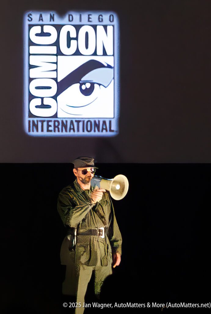 A person in a military-style outfit stands on stage with a megaphone in front of a projected San Diego Comic-Con International logo.
