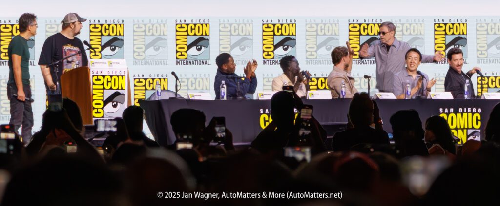 A group of people sit and stand behind a long table on stage at San Diego Comic-Con, with Comic-Con banners in the background and audience members taking photos.