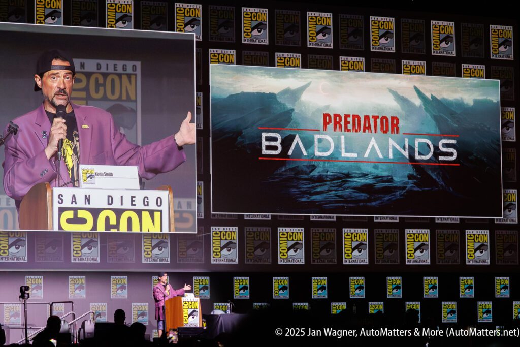 A speaker at San Diego Comic-Con presents "Predator: Badlands" on a large screen, with Comic-Con logos in the background.