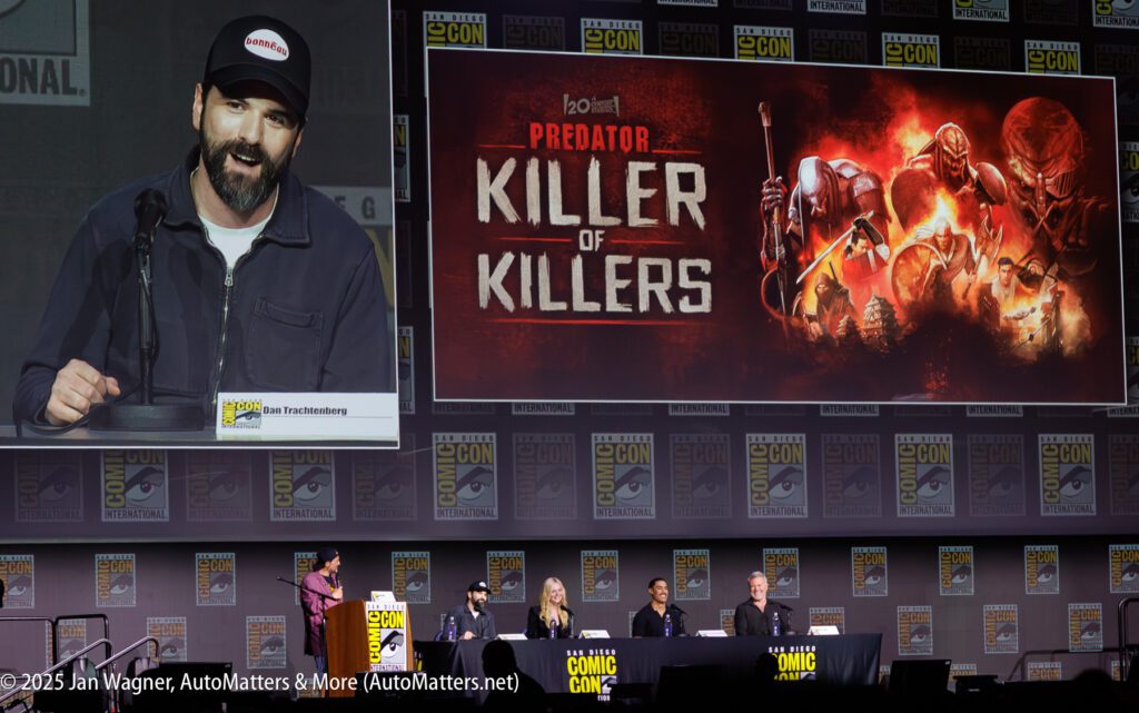 A panel of five people sits at a table onstage at San Diego Comic-Con, with a large "Predator: Killer of Killers" promotional screen and a speaker at a podium.
