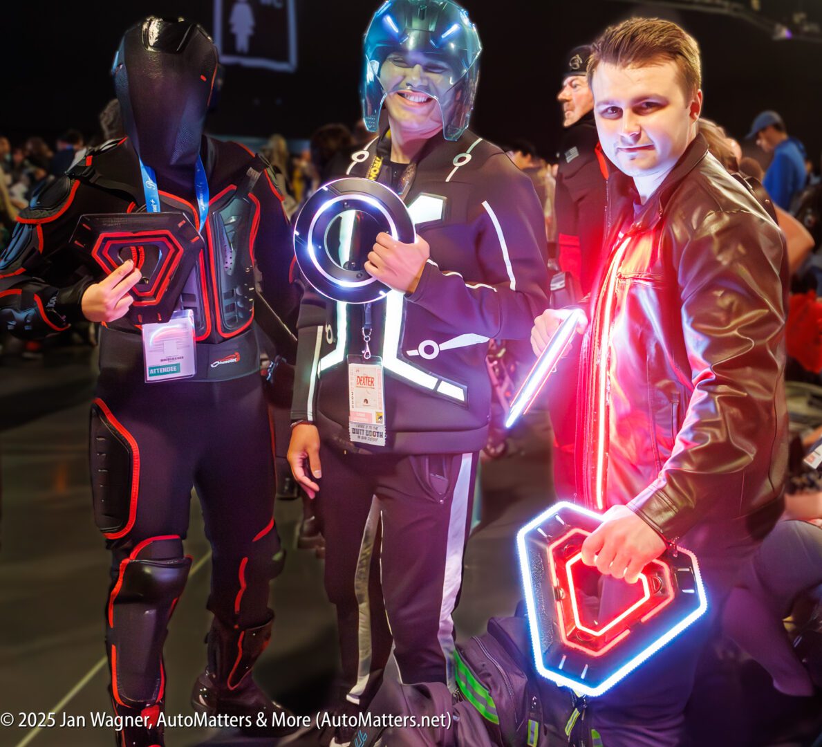 Three people dressed in futuristic costumes with illuminated props pose indoors at an event. Each holds a glowing accessory, and two wear helmets. Crowds are visible in the background.