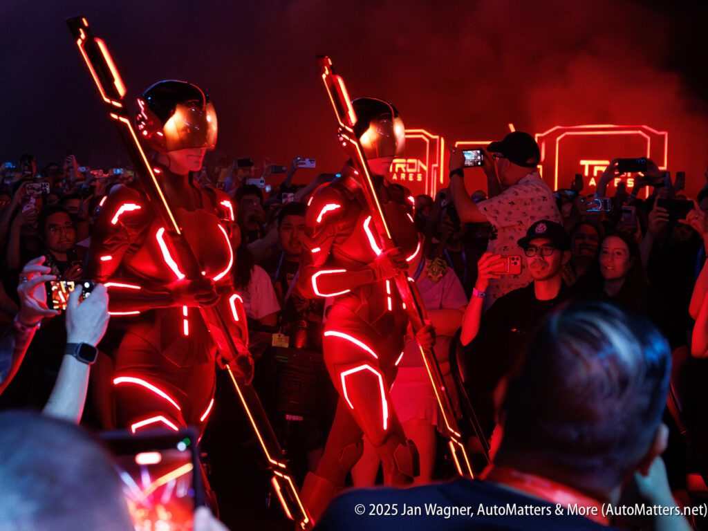 Two individuals in glowing, futuristic suits and helmets walk through a crowd of people taking photos at an event with red lighting.