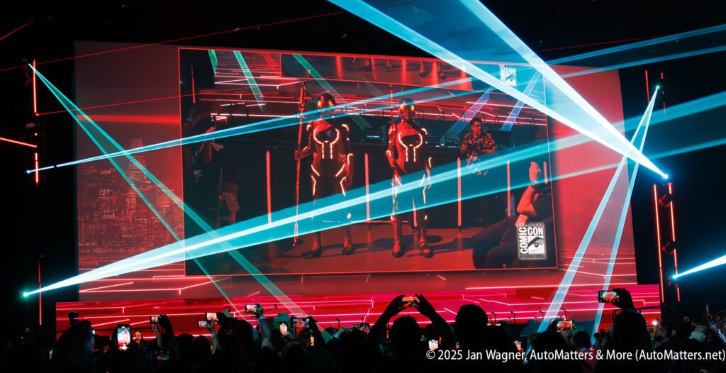 A large screen displays people in costume on stage at a convention, with red and blue laser lights and an audience taking photos.
