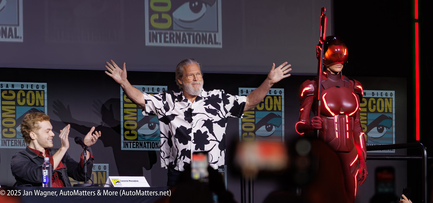 A man stands with arms raised on stage at Comic-Con International, with one person applauding at a table and another in a red futuristic costume and helmet.
