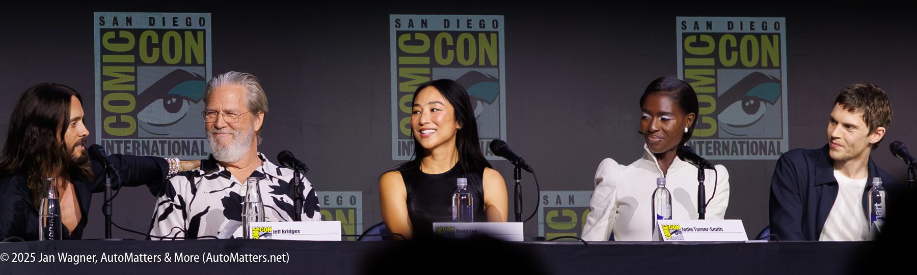 Four panelists sit at a table speaking at San Diego Comic-Con International, with Comic-Con logos displayed on the backdrop behind them.