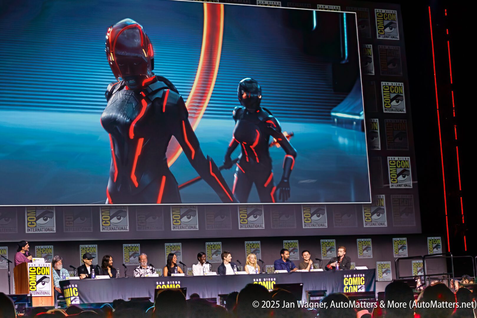 A panel of speakers sits onstage at Comic-Con below a large screen displaying two futuristic, armored characters with glowing red lines.
