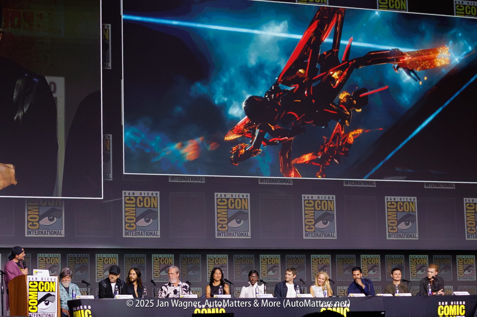 A panel of speakers sits at a long table at Comic-Con, with a large screen behind them displaying a red futuristic aircraft in flight.