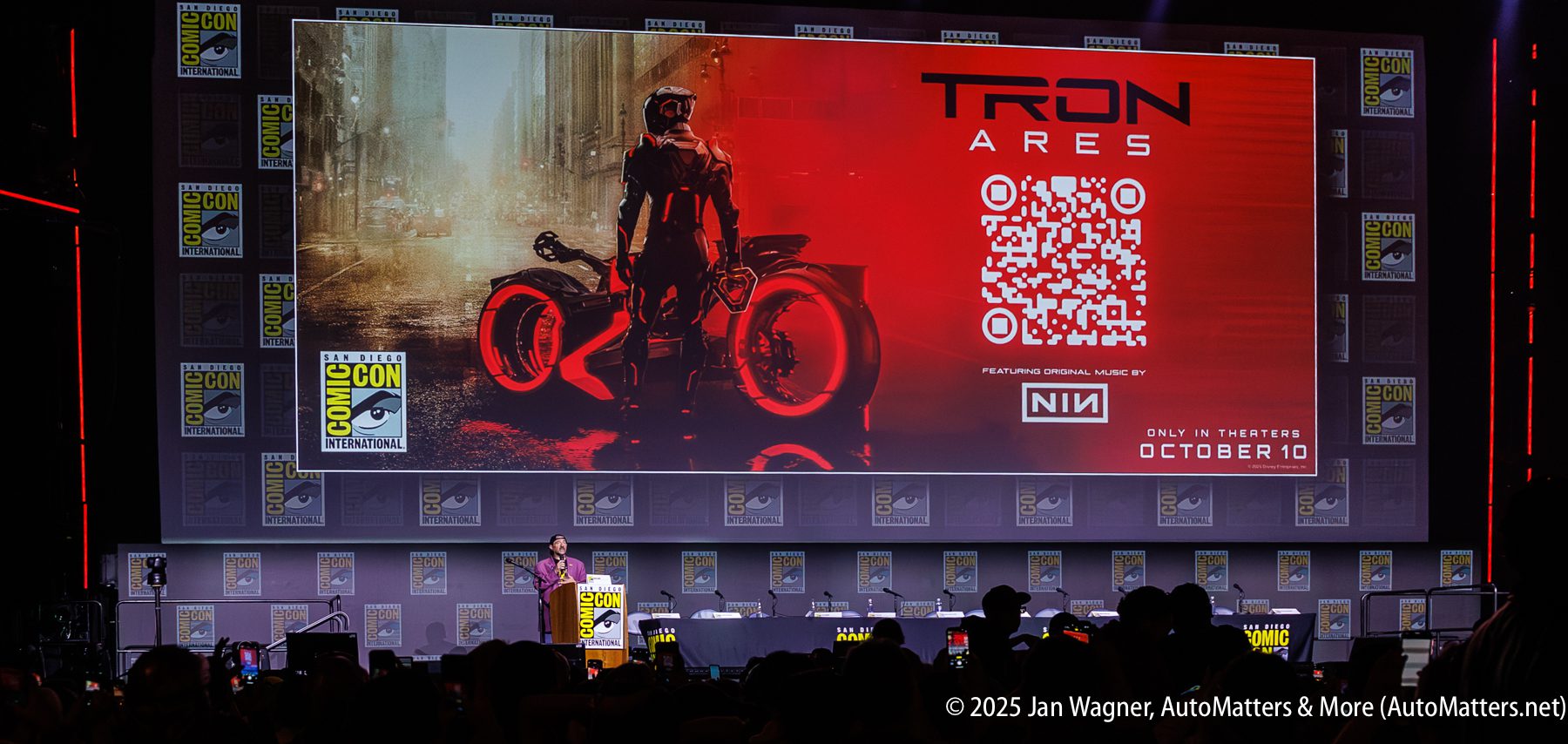 A presenter stands on stage at San Diego Comic-Con before a large screen displaying a "TRON: ARES" movie poster with QR code, release date, and promotional details.