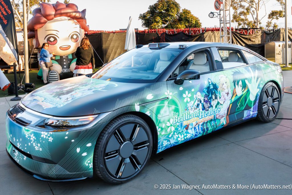 A sedan decorated with anime-themed graphics is parked outdoors near a large anime character inflatable and event tents.