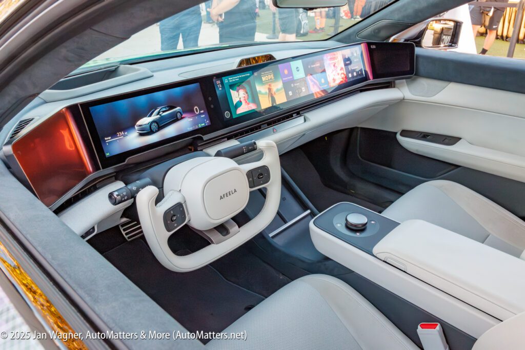 The interior of a modern electric car features a white steering wheel, panoramic dashboard screens, minimalist controls, and light gray seats.