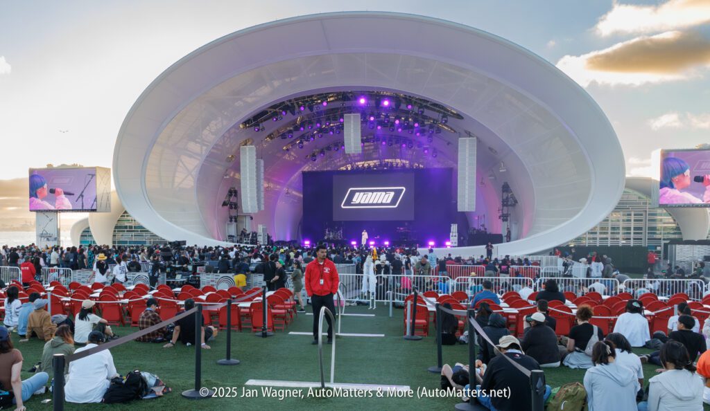 Outdoor concert venue with a large white domed stage, purple lighting, audience seating, and people gathering. Two large screens display the stage event on each side.