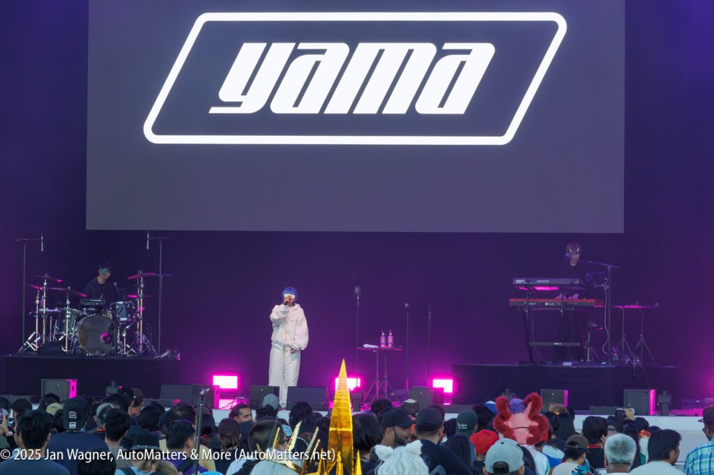 A performer stands on stage in front of a large "yama" logo, with a drummer and keyboardist on either side and an audience watching.