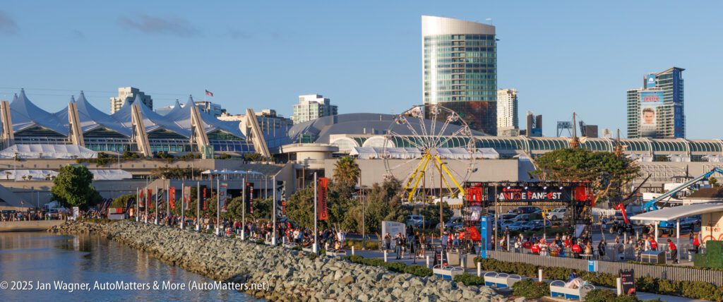 A waterfront scene shows a crowd at an outdoor event near a Ferris wheel, with modern buildings and a convention center in the background under a clear sky.