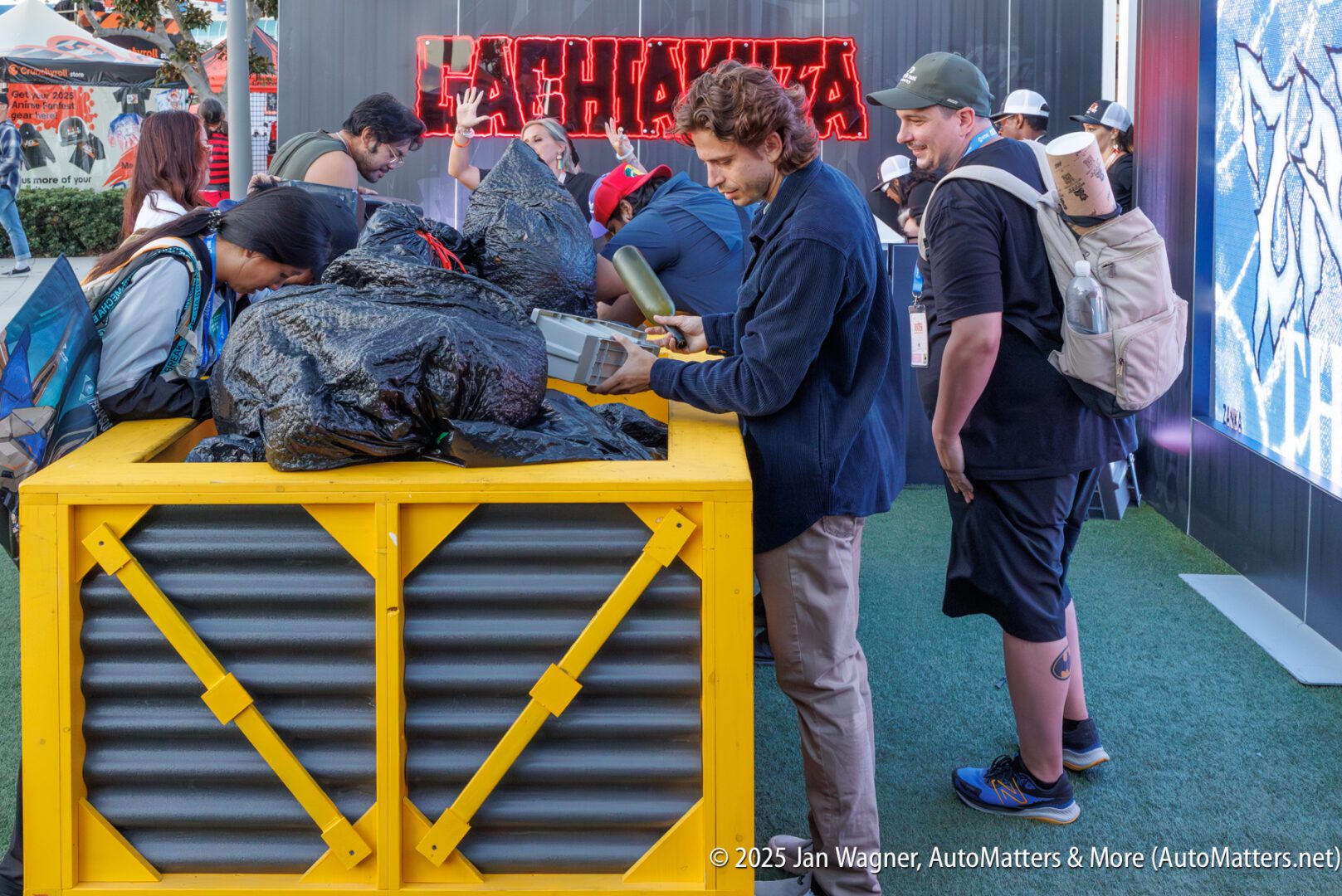People examine a large yellow crate filled with black bags at an outdoor event; others stand nearby, under a red "YAKUZA" sign.