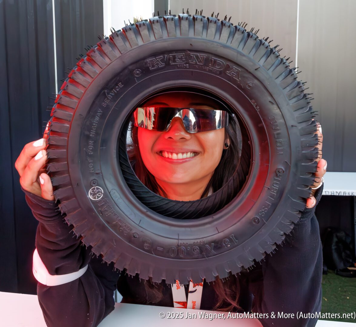 A smiling person wearing sunglasses holds a large tire upright, framing their face within the center opening.