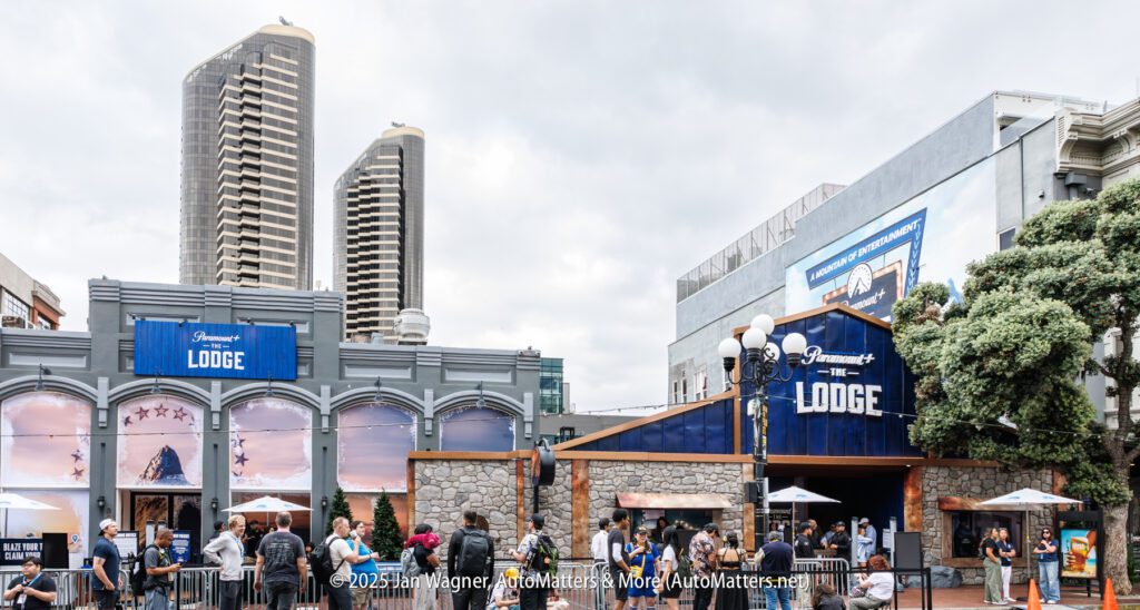 People gather outside the Fairmont Lodge, a themed event space with blue signage, stone accents, and nearby tall buildings under a cloudy sky.