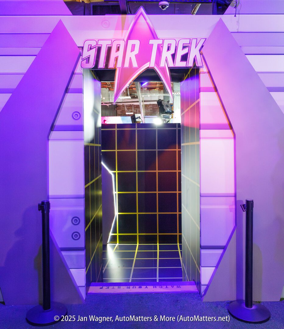 A doorway shaped like the Starfleet insignia with "Star Trek" sign above, flanked by stanchions, leads into a room with a grid pattern on the floor and walls.