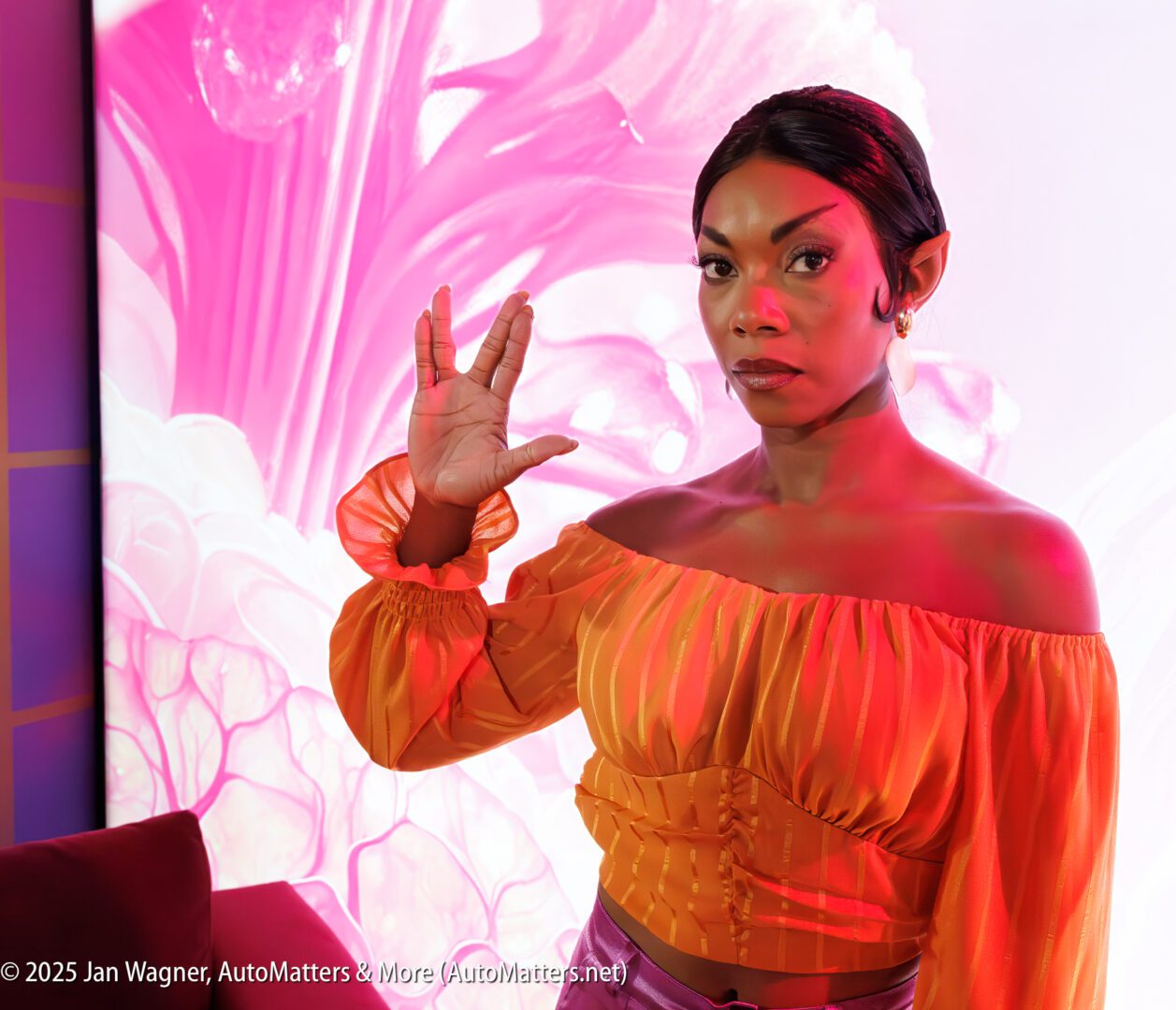 A woman dressed as a Star Trek Vulcan character stands in front of a pink and white backdrop, making the Vulcan salute with her right hand.