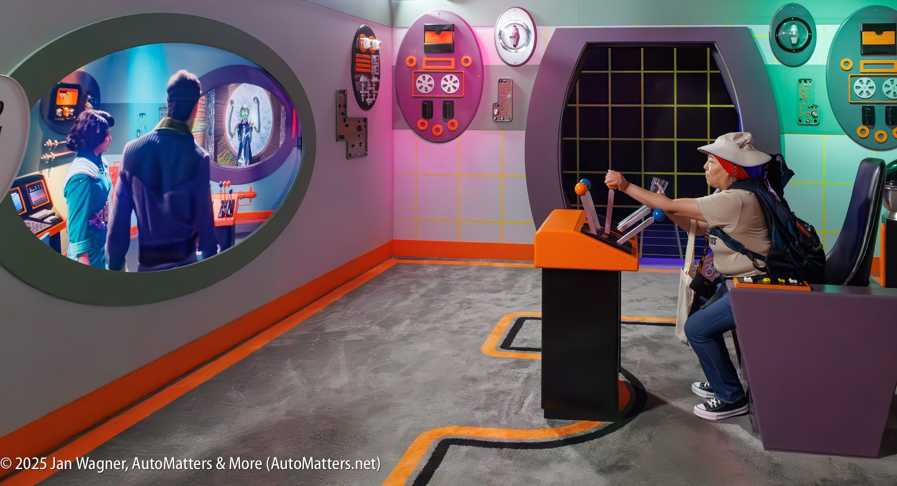 A person sits at a control panel, operating levers while looking at two people displayed on a large screen in a colorful, retro-futuristic room.