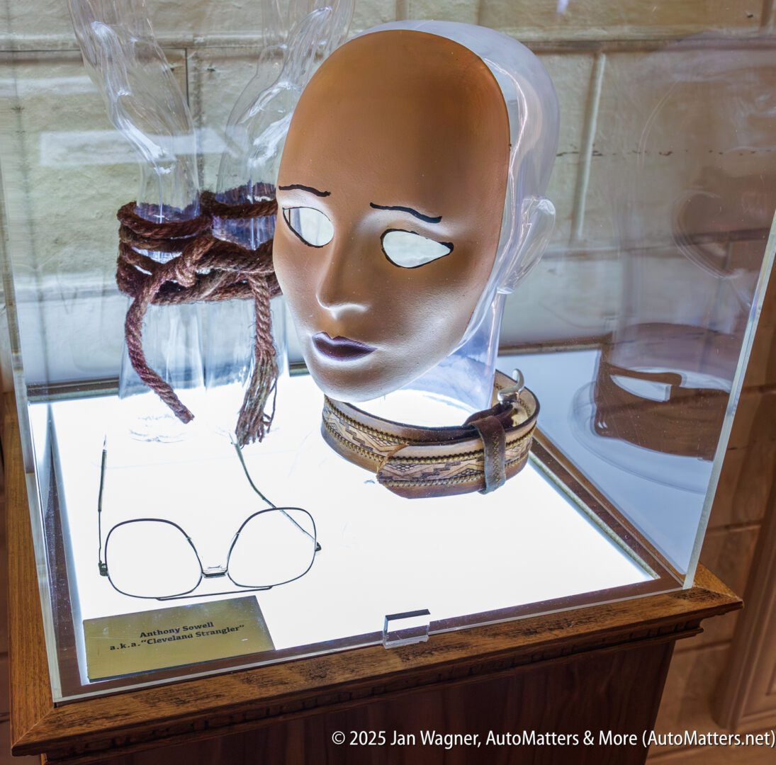 A display case contains a mask, rope, glasses, and a belt, all arranged on a lit surface with a clear cover.