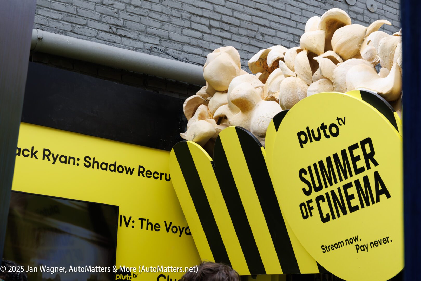 Large popcorn sculpture decorates a yellow and black "SUMMER OF CINEMA" Pluto TV event booth, set against a brick wall.