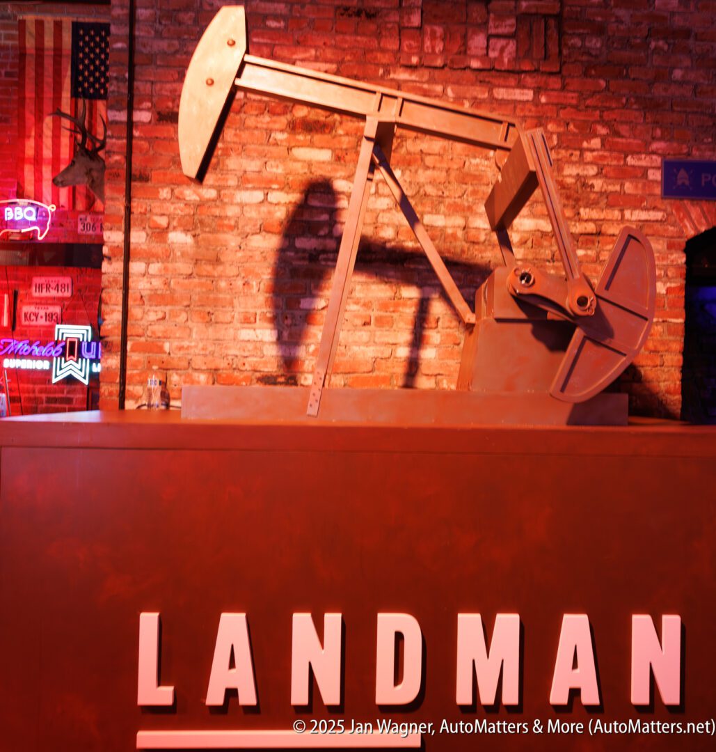 A metal oil pumpjack model is displayed indoors against a brick wall with neon signs, above a counter labeled "LANDMAN.
