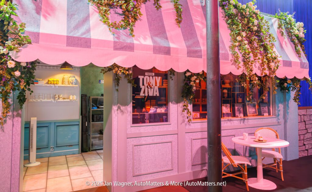 A small café with pink-striped awning, floral decor, and a table with two chairs outside. Shelves and kitchen equipment are visible through the window.