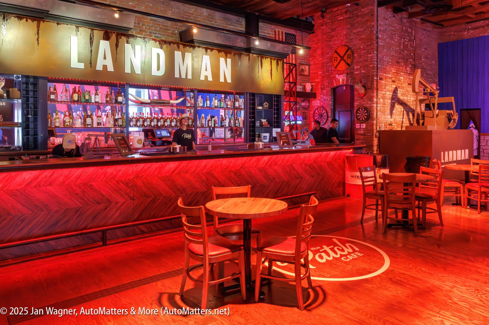 A modern bar and restaurant interior with wooden tables and chairs, a well-lit bar area, and a large "LANDMAN" sign above the counter.