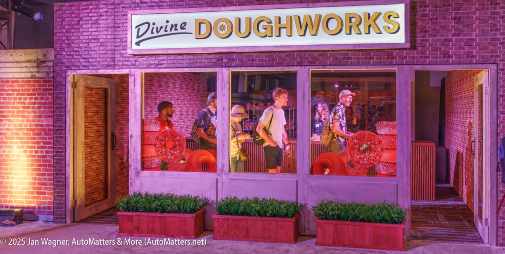 Several people stand in line inside a brick-walled doughnut shop called "Divine Doughworks," holding large decorative red doughnuts.