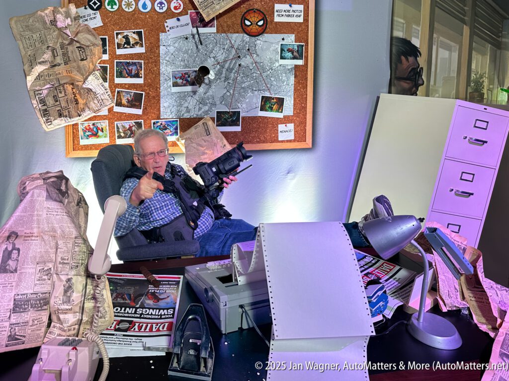 An older man sits in a cluttered office set filled with newspapers, a typewriter, filing cabinets, and a bulletin board covered in photos and Spider-Man memorabilia.