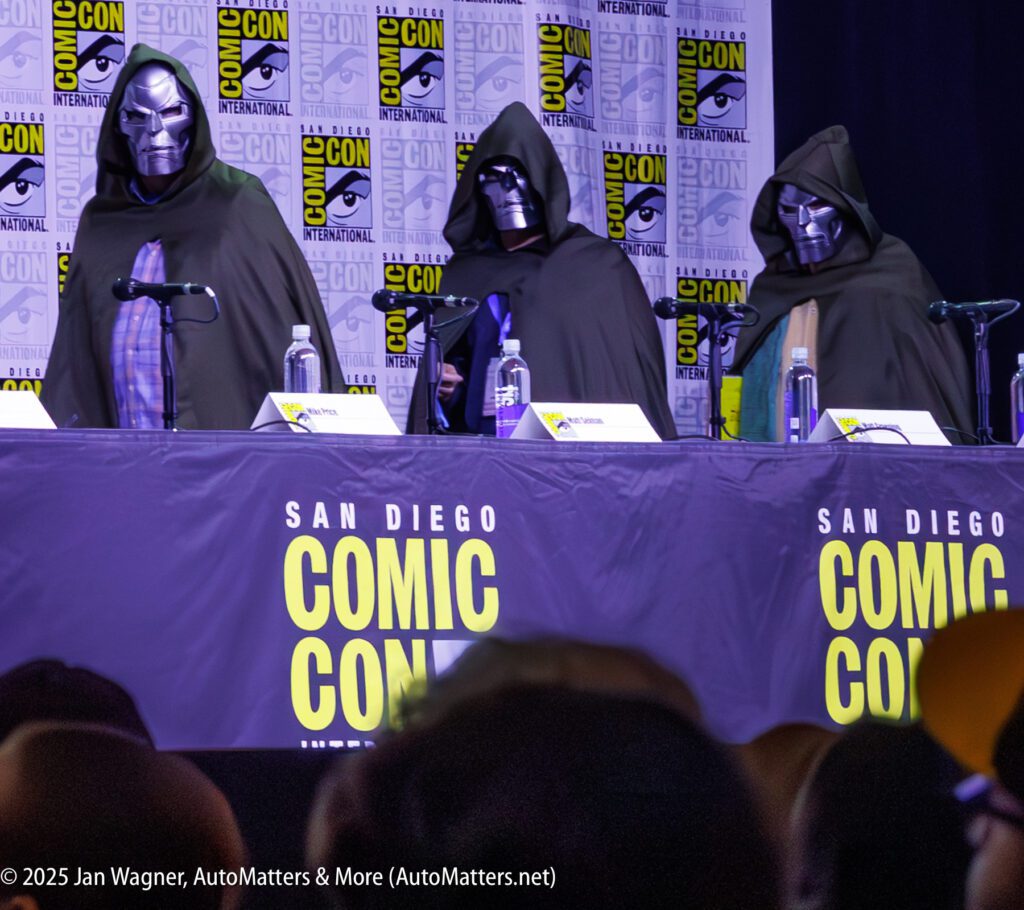 Three people wearing Doctor Doom costumes sit behind a table at a San Diego Comic-Con panel, with Comic-Con logos visible on the backdrop and table skirt.