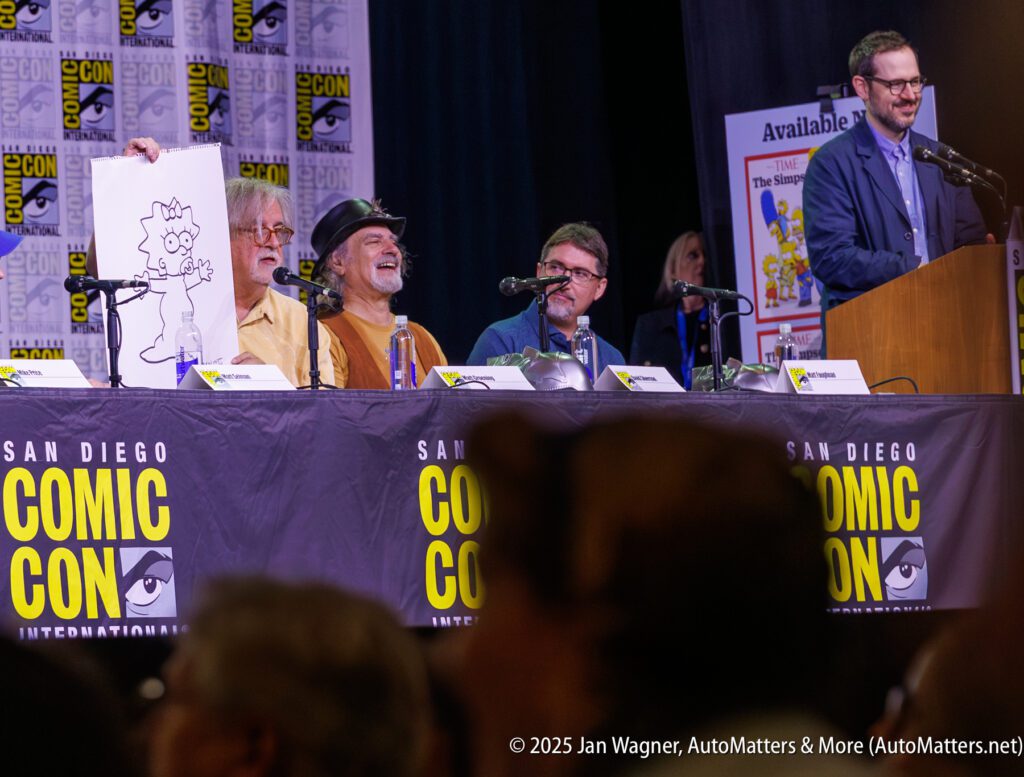 Four panelists sit at a San Diego Comic-Con table; one holds up a drawing of Lisa Simpson as a cat while another person stands at a podium.