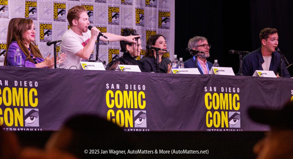 Six panelists speak and interact on stage at San Diego Comic-Con, seated behind a branded table with microphones and name cards.