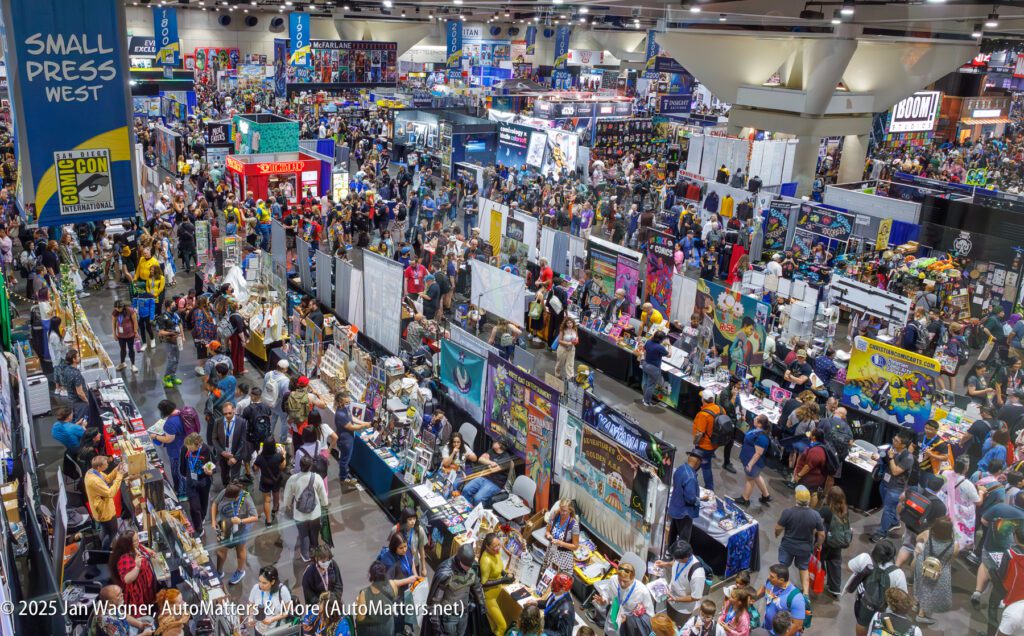 Large crowd attending a busy comic convention in a spacious exhibition hall, with booths, banners, and merchandise displays filling the venue.