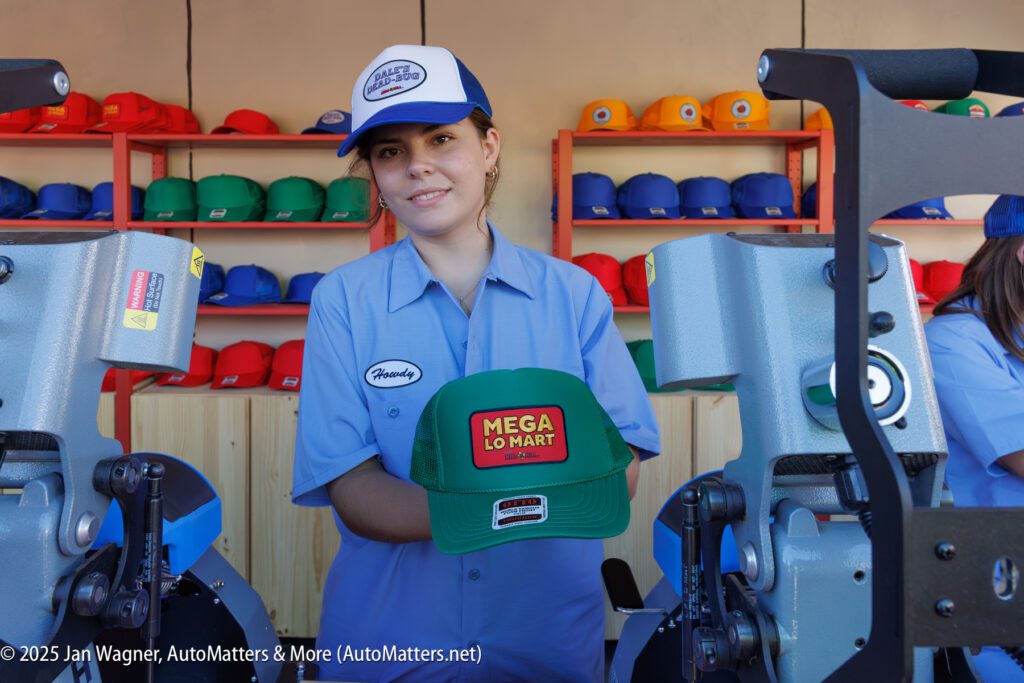 A person wearing a blue uniform and cap holds a green "Mega Lo Mart" hat in front of hat printing machines, with colorful hats displayed on shelves behind.
