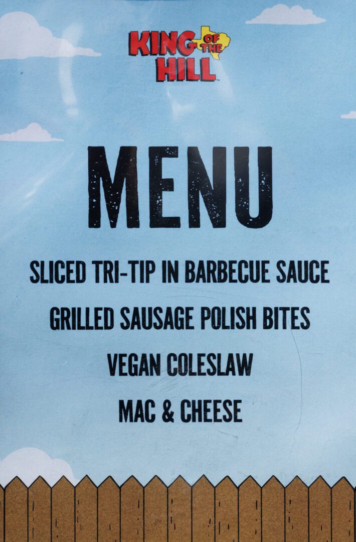 A printed "King of the Hill" menu lists sliced tri-tip in barbecue sauce, grilled sausage Polish bites, vegan coleslaw, and mac & cheese on a blue background.