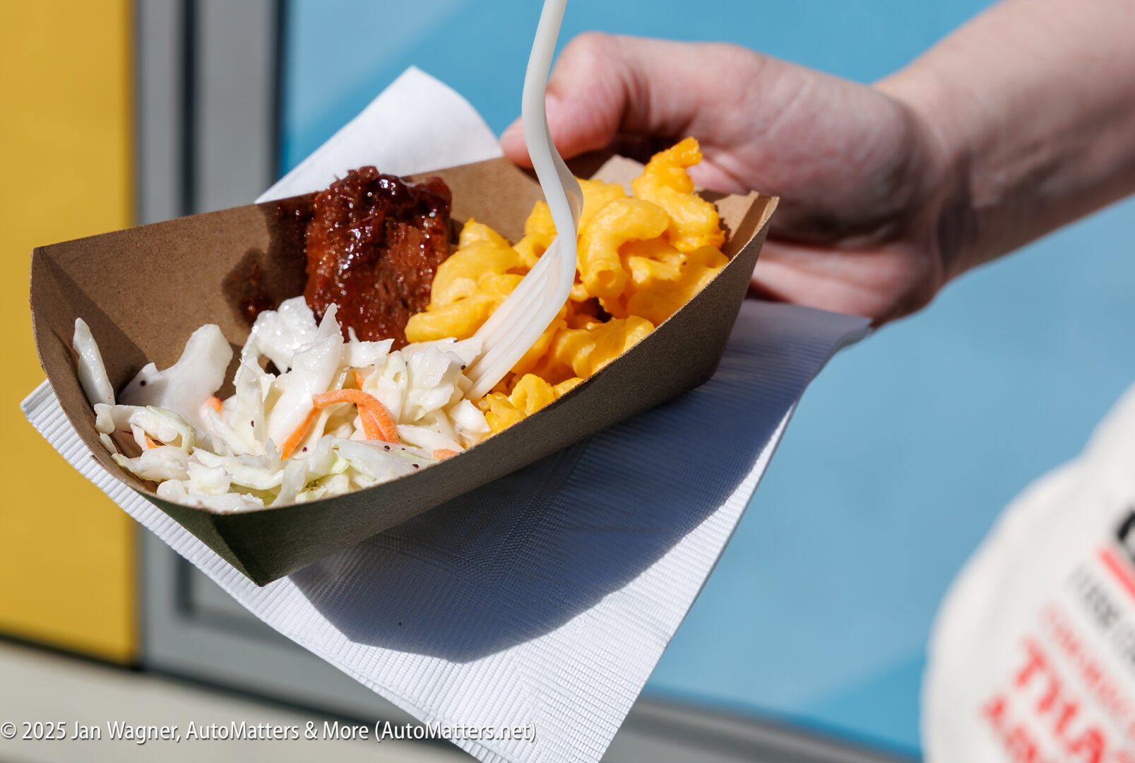 A hand holds a cardboard tray with macaroni and cheese, coleslaw, barbecue meat, and a plastic fork, resting on a napkin.