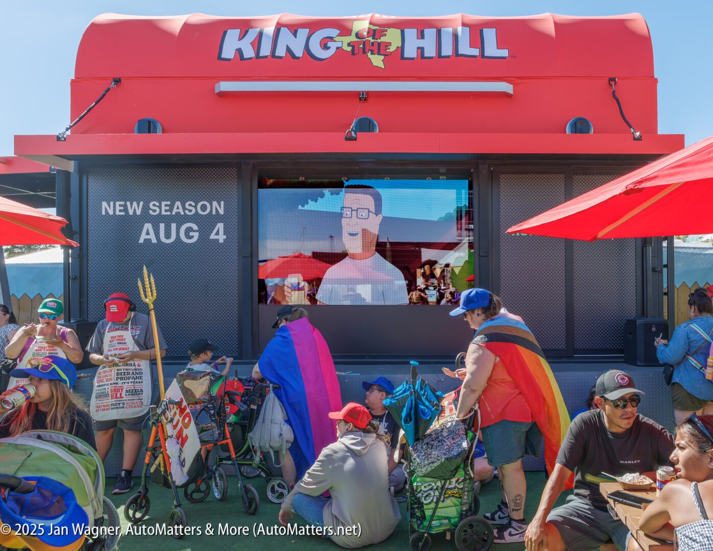 A crowd gathers under red umbrellas in front of a "King of the Hill" display advertising a new season starting August 4. A screen shows an animated character from the show.