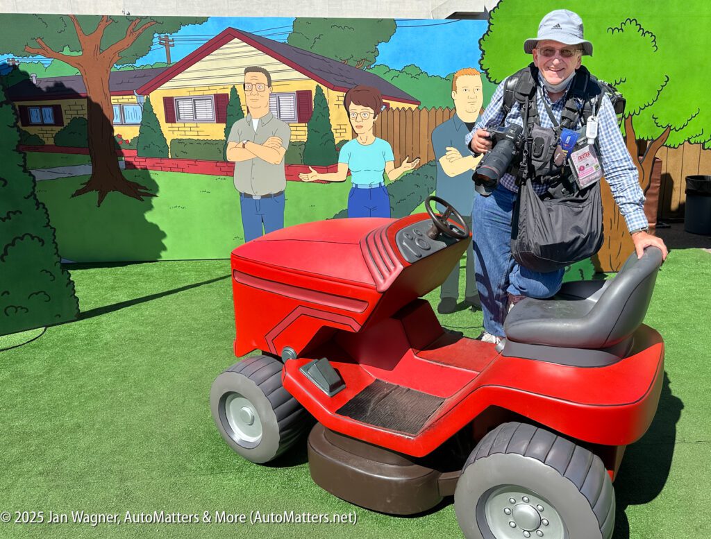 A person poses with one foot on a large red lawn mower, standing in front of a cartoon mural depicting three people in a suburban backyard.