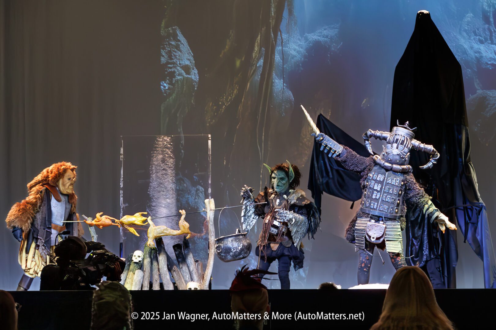 Three people in elaborate fantasy costumes perform on stage, featuring a lion-like figure, a green-skinned character, and a large armored figure with a sword, with dramatic lighting and props.