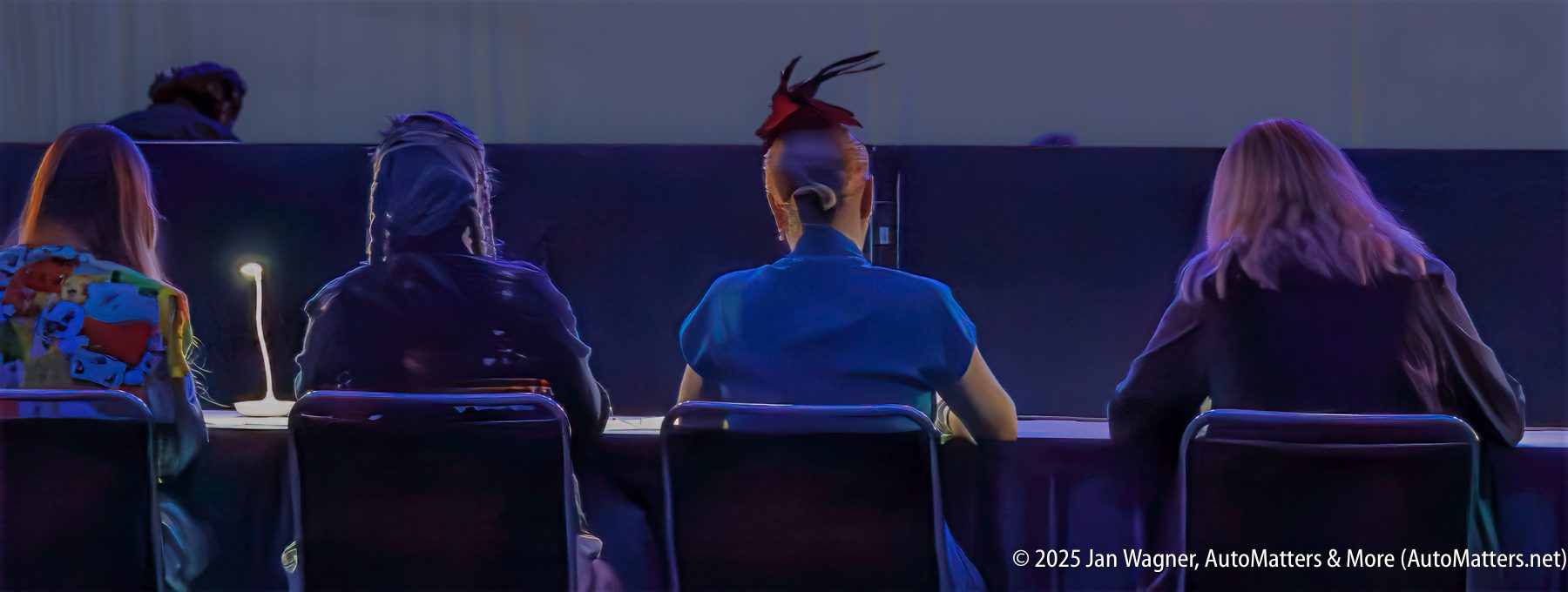 Four people sit at a table facing a stage, viewed from behind; one has a colorful backpack, another has a feathered headpiece.