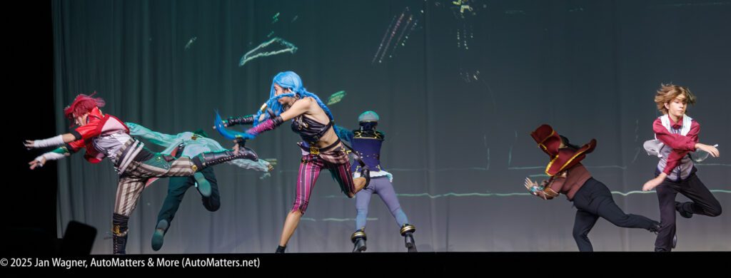 Five people in colorful, elaborate costumes perform a dynamic action pose on stage, with three kicking and two in mid-motion, against a plain backdrop.